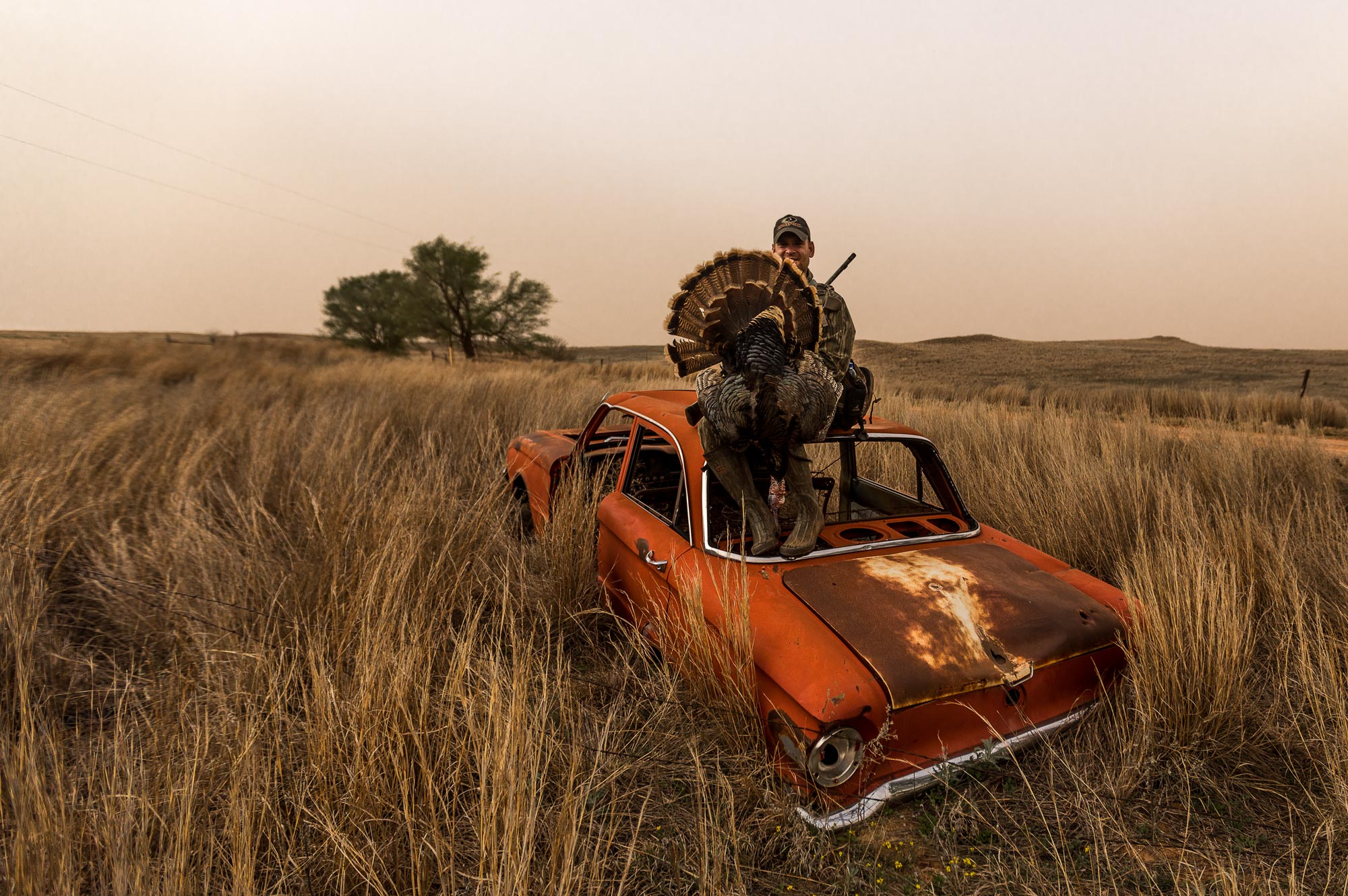 turkey hunter posing with a turkey on the hood of an old car in a field