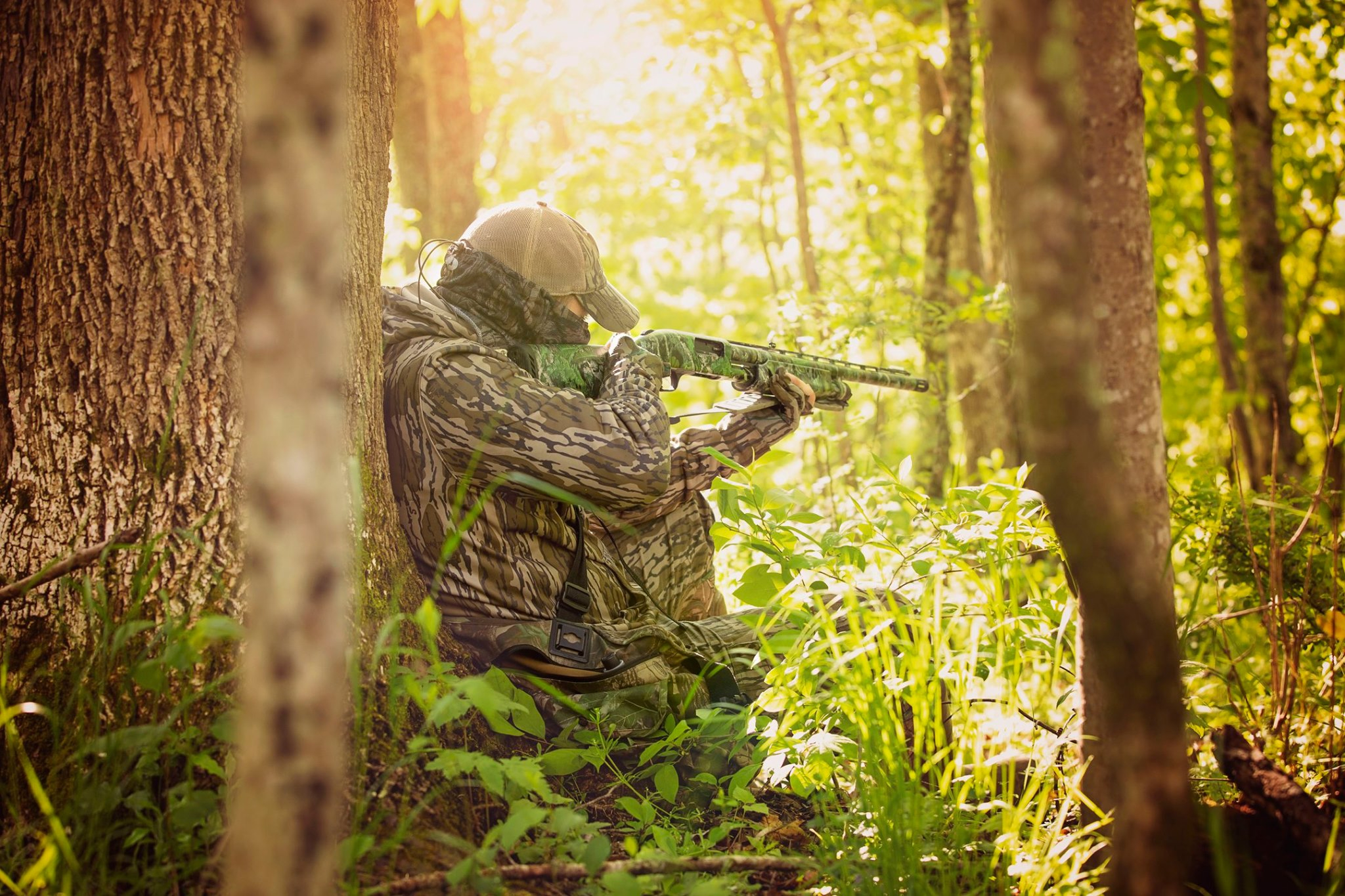 A hunter, leaning up against a tree, takes aim.